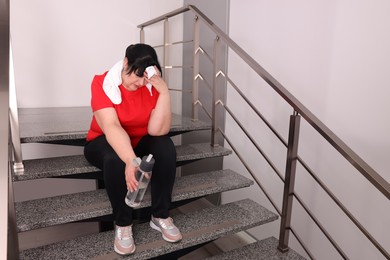 Photo of Tired overweight mature woman with towel and bottle of water sitting on stairs indoors, space for text