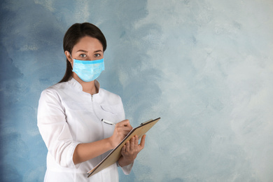 Photo of Doctor with disposable mask on face holding clipboard against light blue background. Space for text