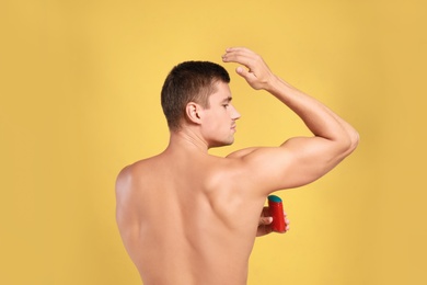 Young man applying deodorant to armpit on yellow background