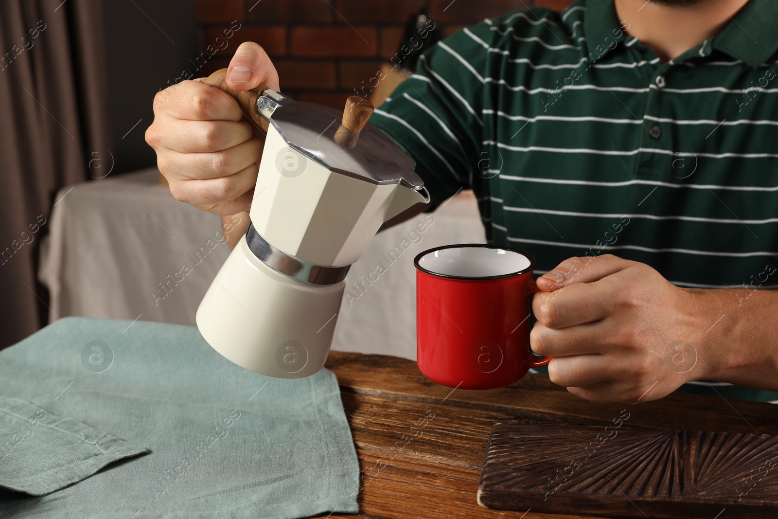 Photo of Man pouring aromatic coffee from moka pot into mug at wooden table in kitchen, closeup