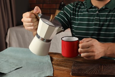 Photo of Man pouring aromatic coffee from moka pot into mug at wooden table in kitchen, closeup