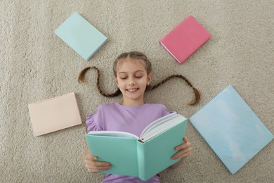 Photo of Cute little girl reading book on floor at home, top view