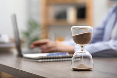 Photo of Hourglass with flowing sand on desk. Woman using laptop indoors, selective focus