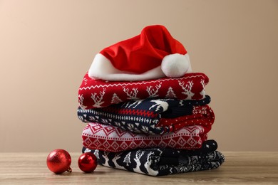 Photo of Stack of different Christmas sweaters, Santa hat and baubles on wooden table against beige background