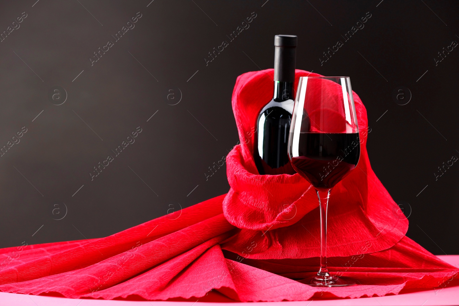 Photo of Stylish presentation of delicious red wine in bottle and glass on pink table against dark background. Space for text