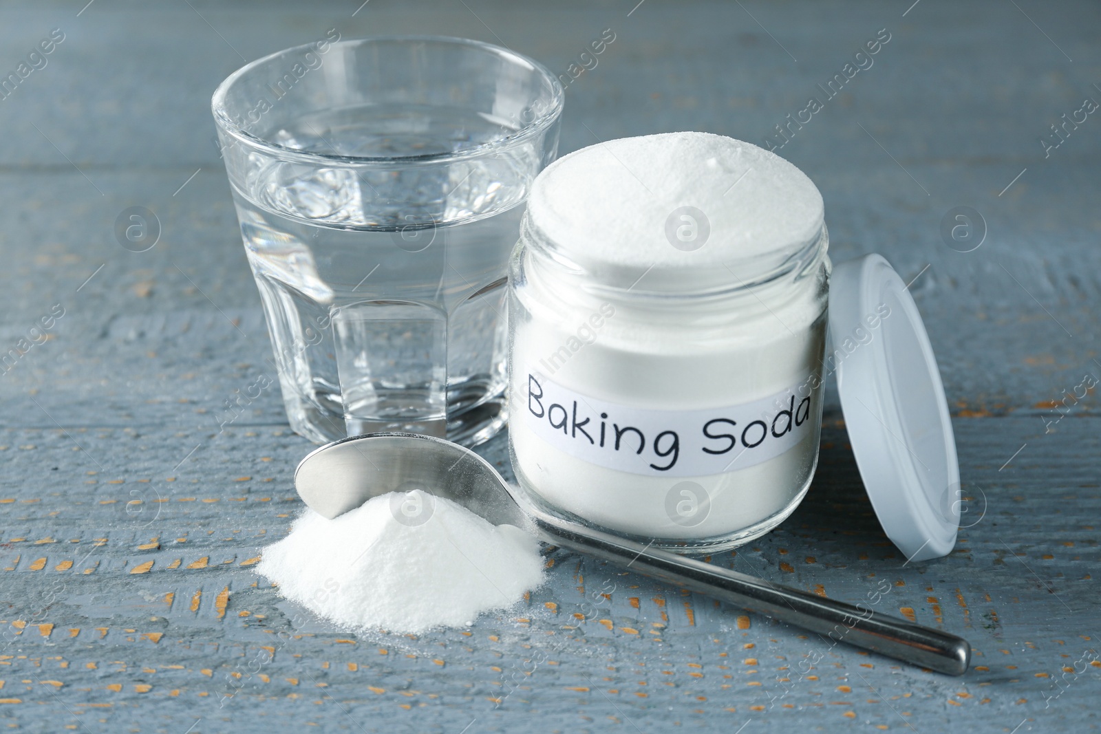 Photo of Jar and spoon with baking soda near glass of water on light blue wooden table, closeup