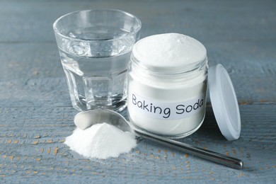 Jar and spoon with baking soda near glass of water on light blue wooden table, closeup