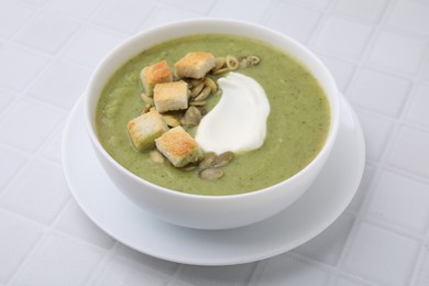 Photo of Delicious broccoli cream soup with croutons, sour cream and pumpkin seeds on white tiled table, closeup