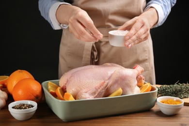 Woman adding salt to raw chicken with orange slices at wooden table, closeup