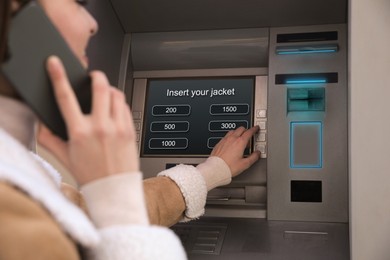 Young woman talking by mobile phone while using cash machine for money withdrawal outdoors, closeup