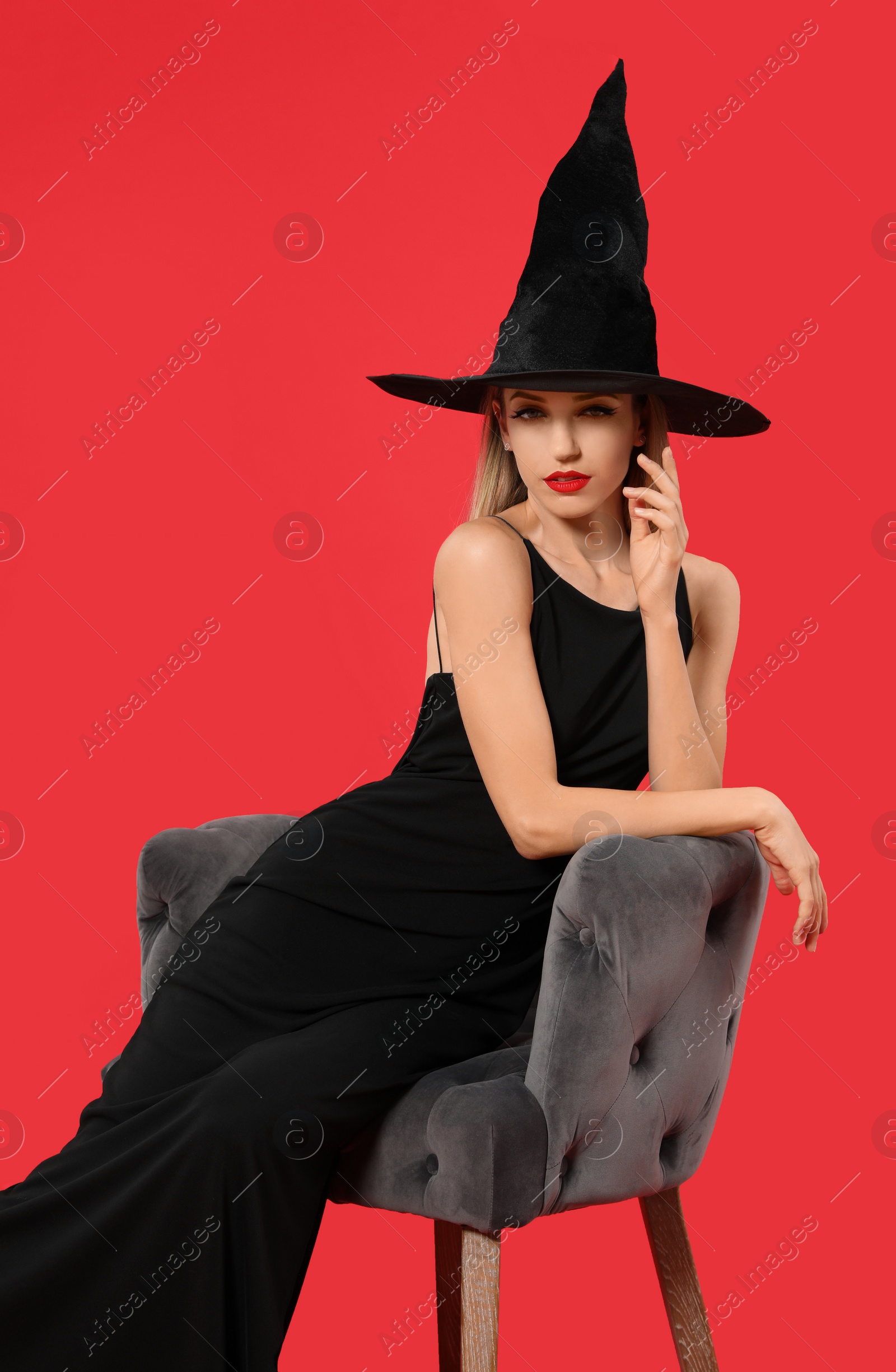 Photo of Beautiful woman wearing witch costume in armchair on red background. Halloween party