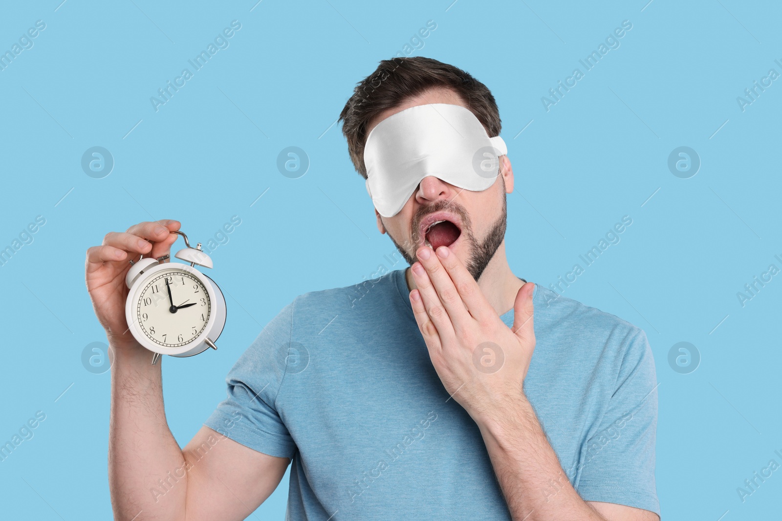 Photo of Tired man with sleep mask and alarm clock on light blue background. Insomnia problem