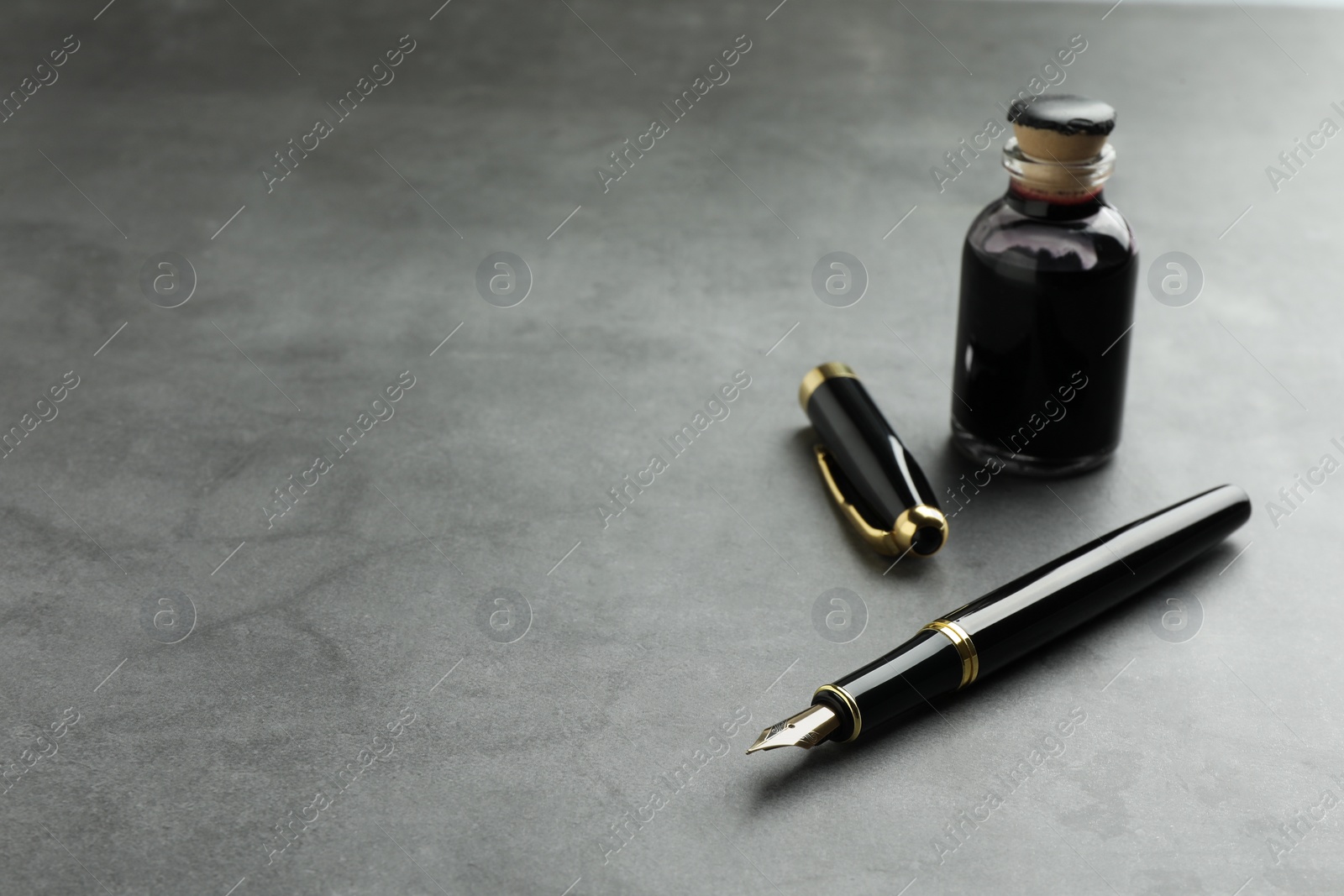 Photo of Stylish fountain pen, bottle of ink and inkwell on grey textured table, space for text