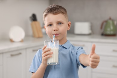 Cute boy drinking fresh milk from glass and showing thumb up indoors