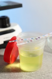 Photo of Container with urine sample for analysis and test strips on grey table in laboratory