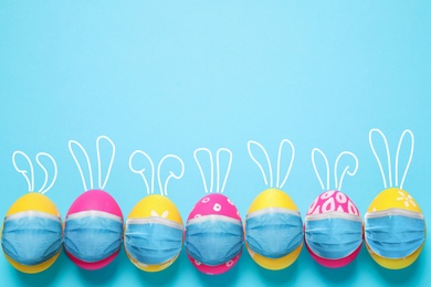 Image of COVID-19 pandemic. Colorful Easter eggs with cute bunny ears in protective masks and space for text on light blue background, flat lay