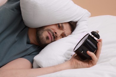 Photo of Sleepy man holding alarm clock and covering his ears with pillows in bed