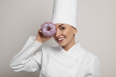 Photo of Happy professional confectioner in uniform holding delicious doughnut on light grey background