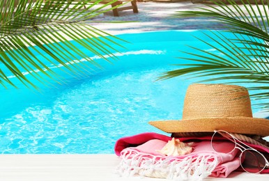 Beach towel, hat and sunglasses on white wooden surface near outdoor swimming pool. Space for text