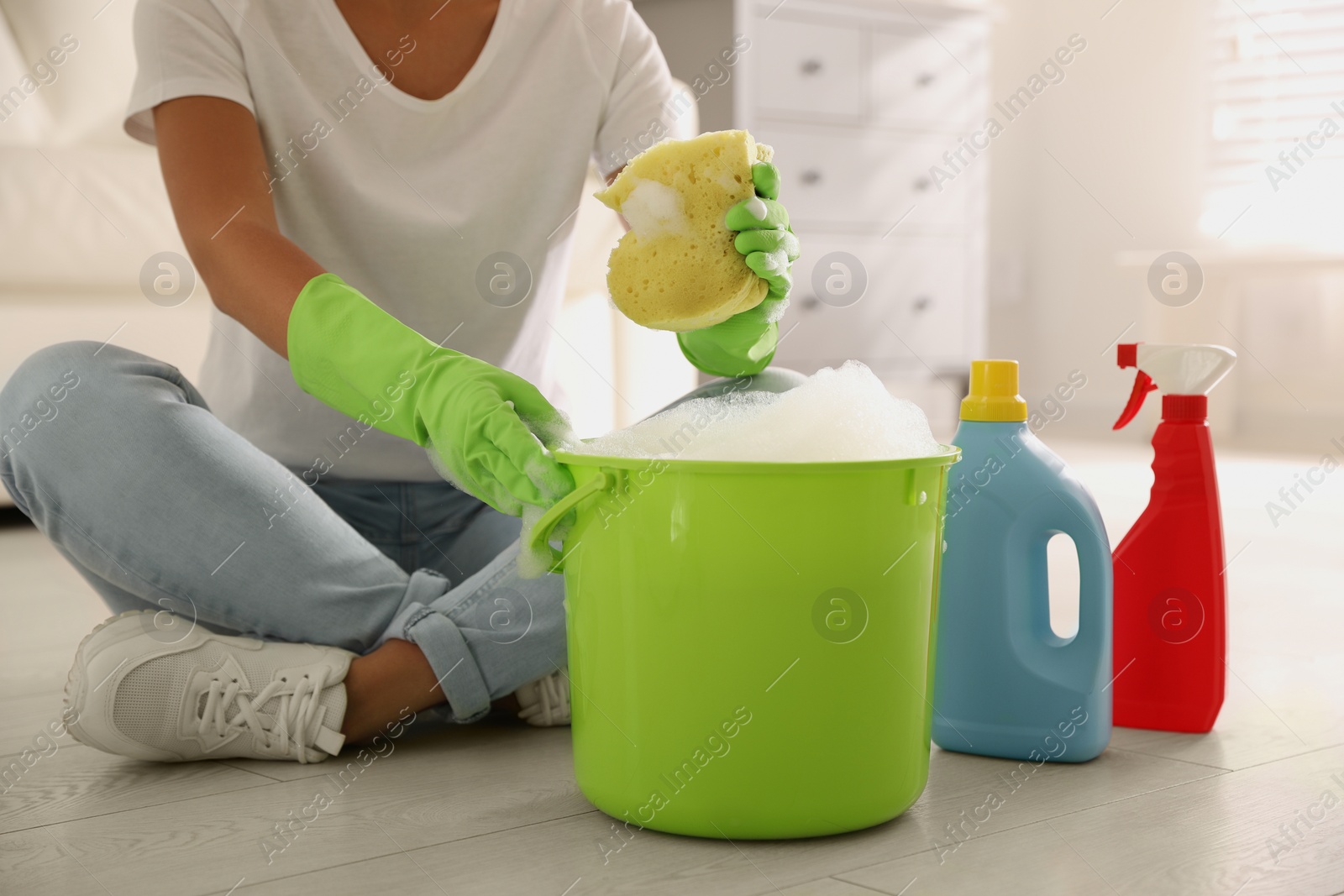 Photo of Woman holding sponge with foam over bucket indoors, closeup. Cleaning supplies