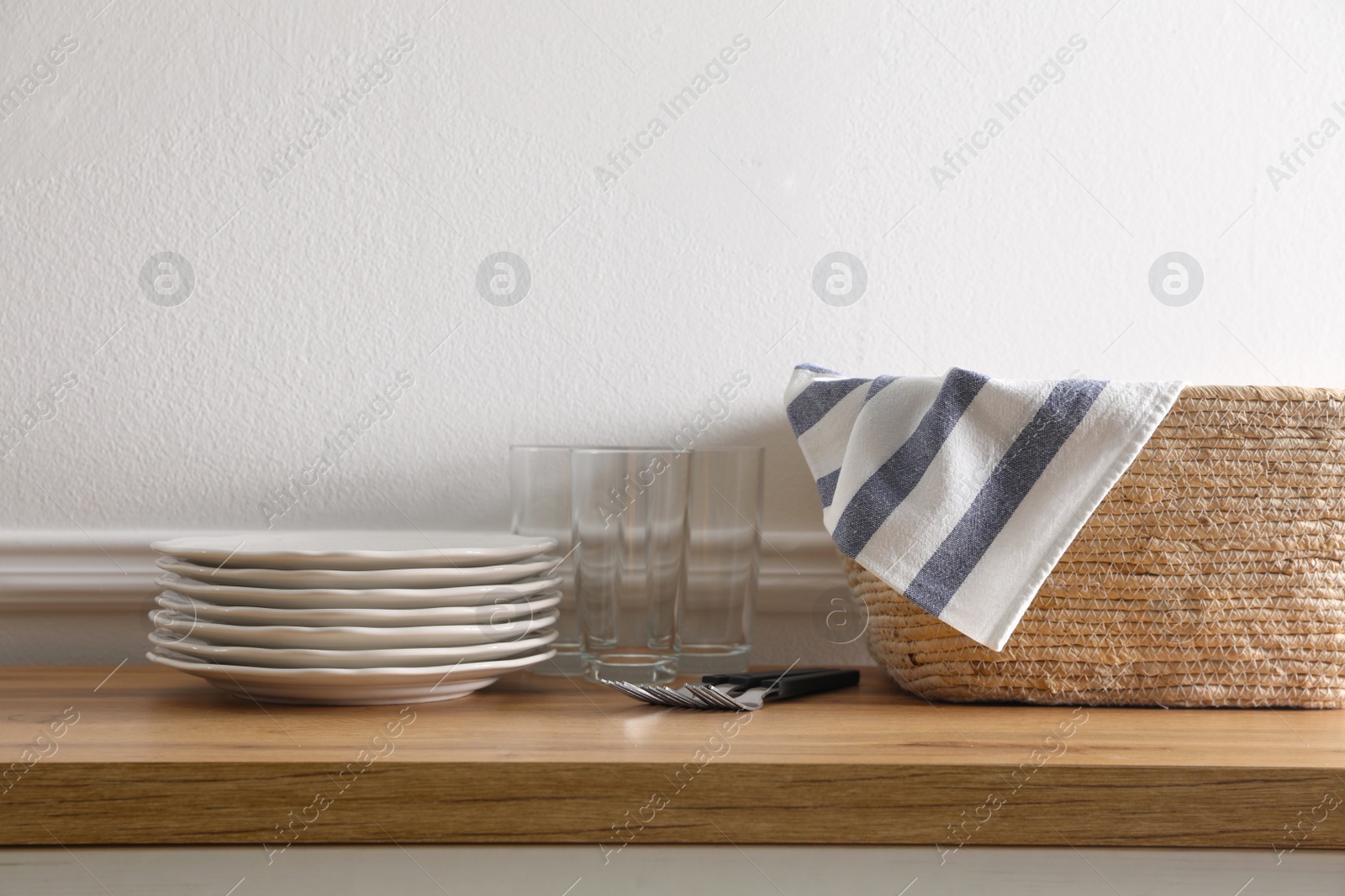 Photo of Kitchen towel in wicker basket and clean dishware on wooden table near white wall