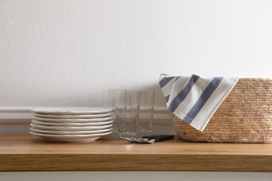 Photo of Kitchen towel in wicker basket and clean dishware on wooden table near white wall