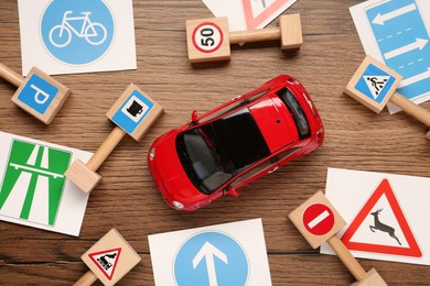 Different road signs and toy car on wooden table, flat lay. Driving school