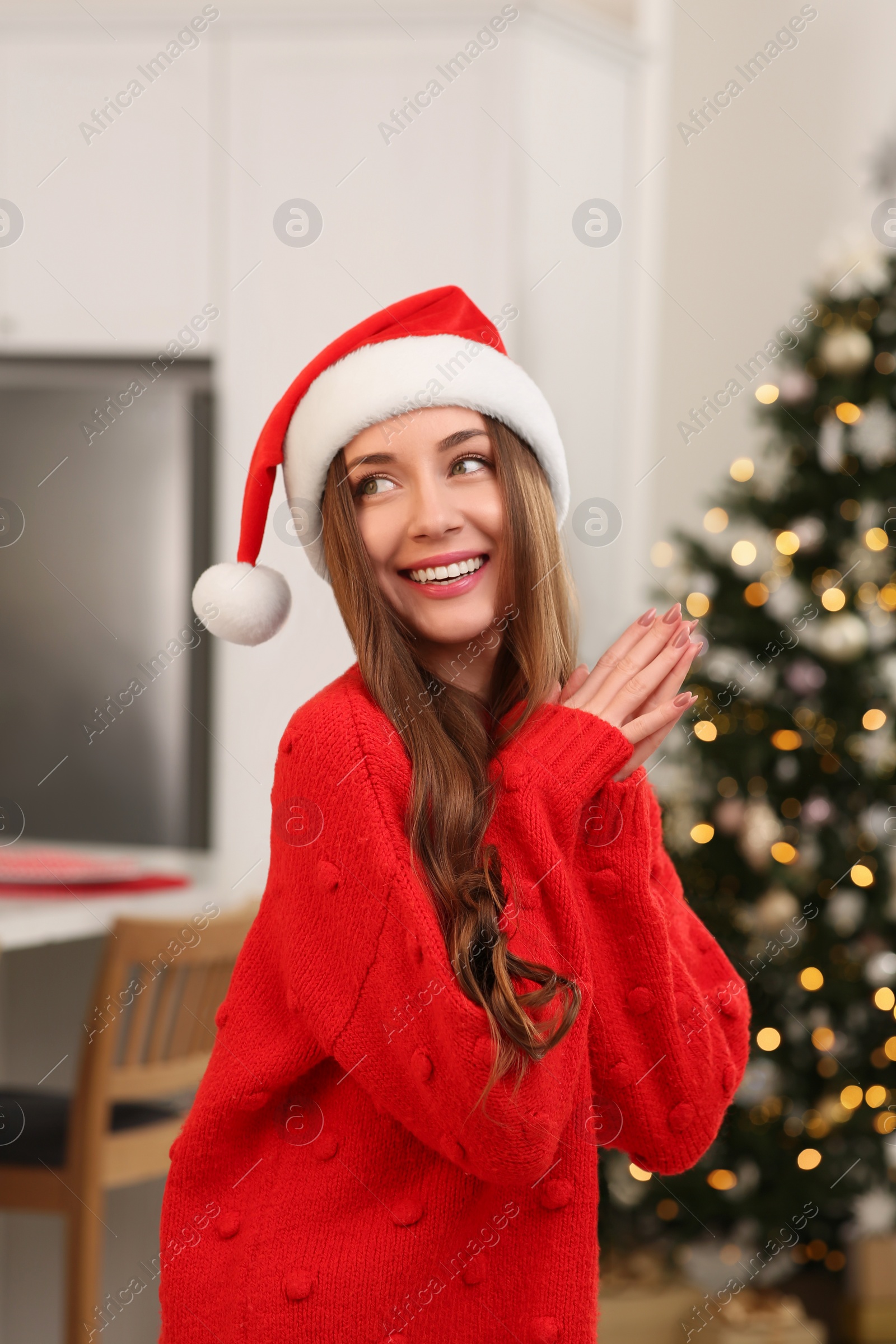 Photo of Beautiful young smiling woman in Santa hat near Christmas tree at home