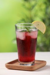 Photo of Refreshing hibiscus tea with ice cubes, mint and lemon in glass on white table against blurred green background