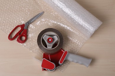 Photo of Roll of bubble wrap, scissors and adhesive tape on wooden table, flat lay