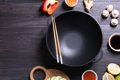 Photo of Empty iron wok, chopsticks and ingredients on dark grey wooden table, flat lay. Space for text