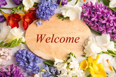 Welcome card. Wooden board with word surrounded by beautiful spring flowers on white background, flat lay