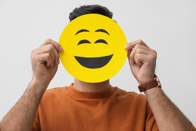 Man hiding emotions using card with drawn smiling face on white background