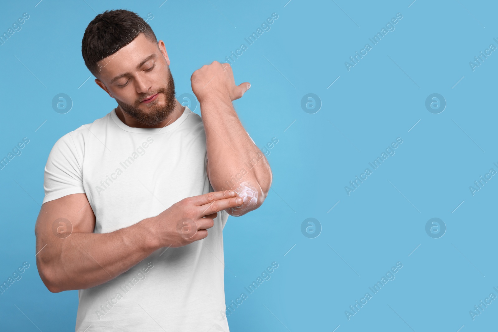 Photo of Handsome man applying body cream onto his elbow on light blue background, space for text