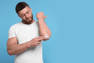 Photo of Handsome man applying body cream onto his elbow on light blue background, space for text