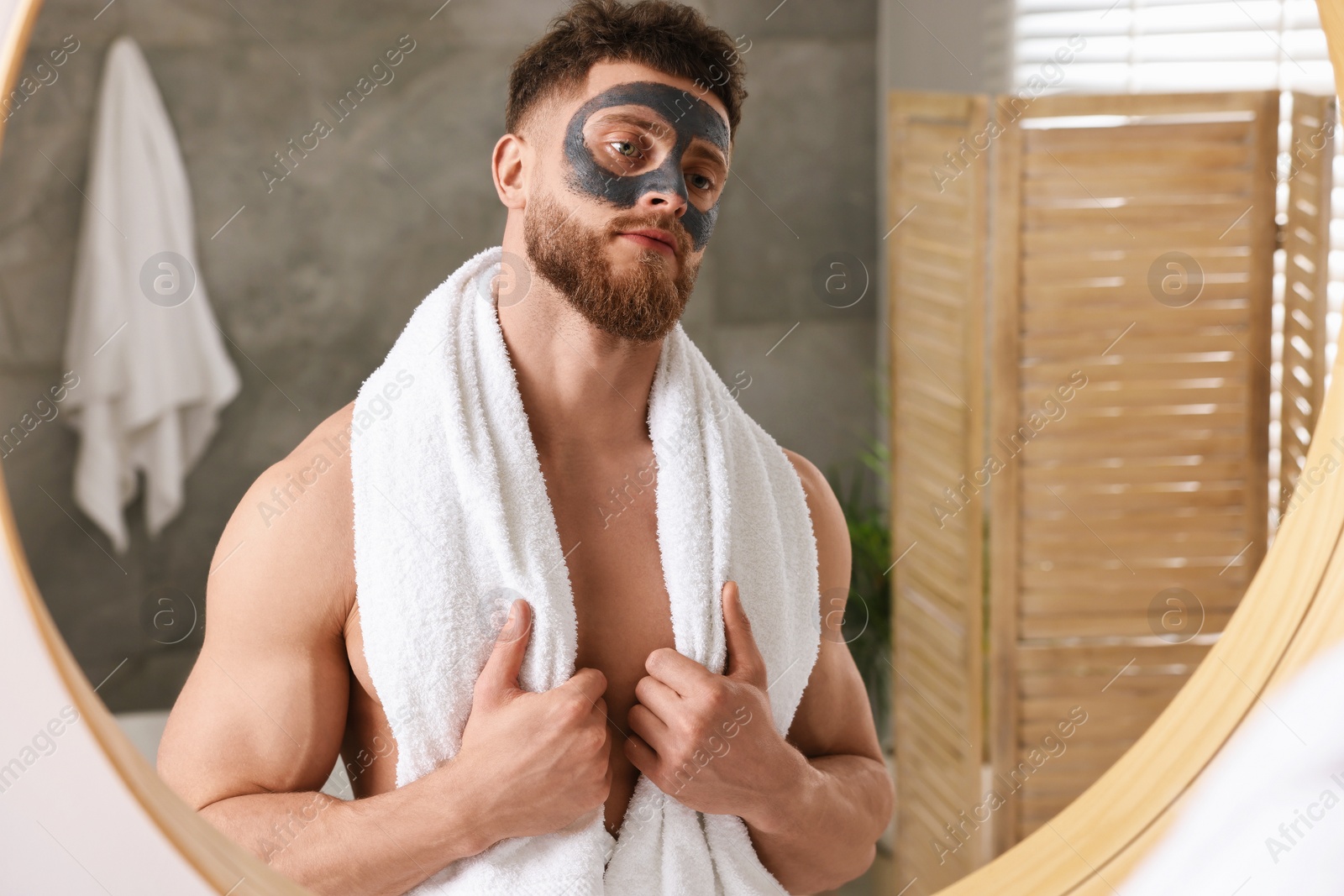 Photo of Handsome man with clay mask on his face near mirror in bathroom