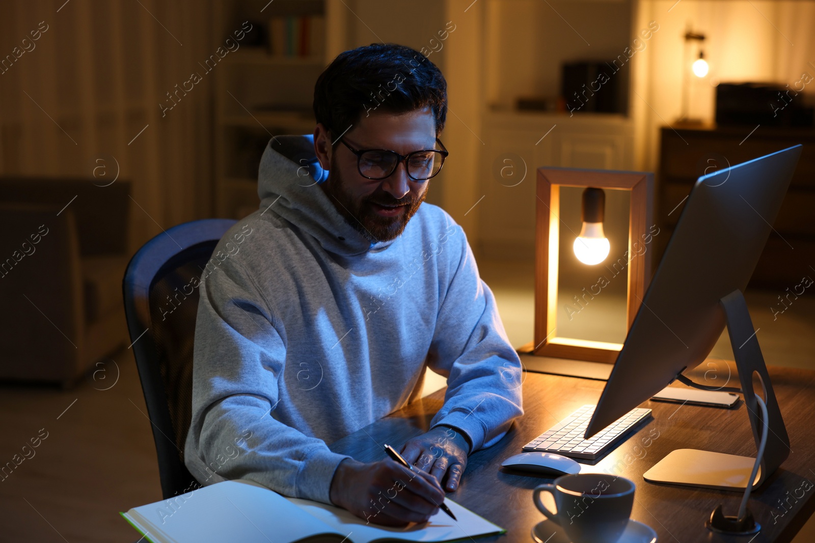 Photo of Home workplace. Man taking notes while working with computer at wooden desk in room at night