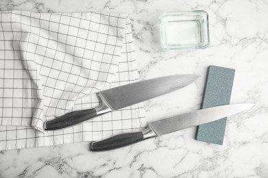 Photo of Sharpening stone, knives and water on white marble table, flat lay