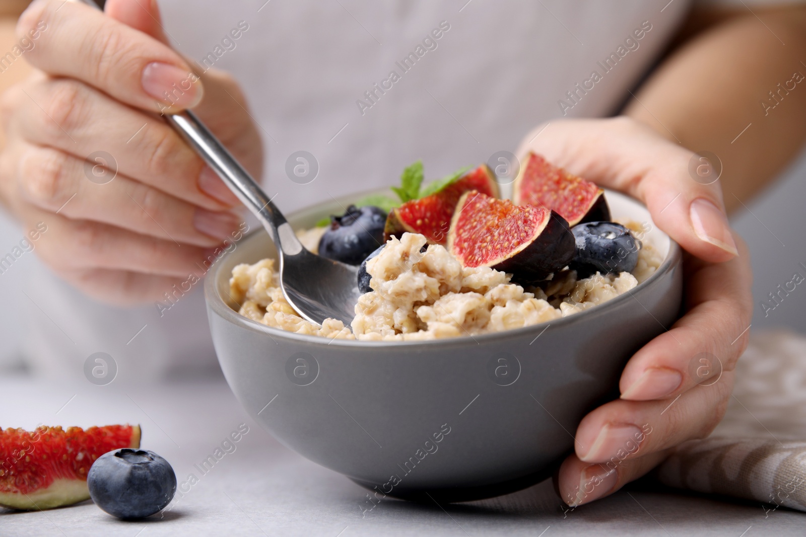 Photo of Woman holding bowl of oatmeal with blueberries and fig pieces at white table, closeup