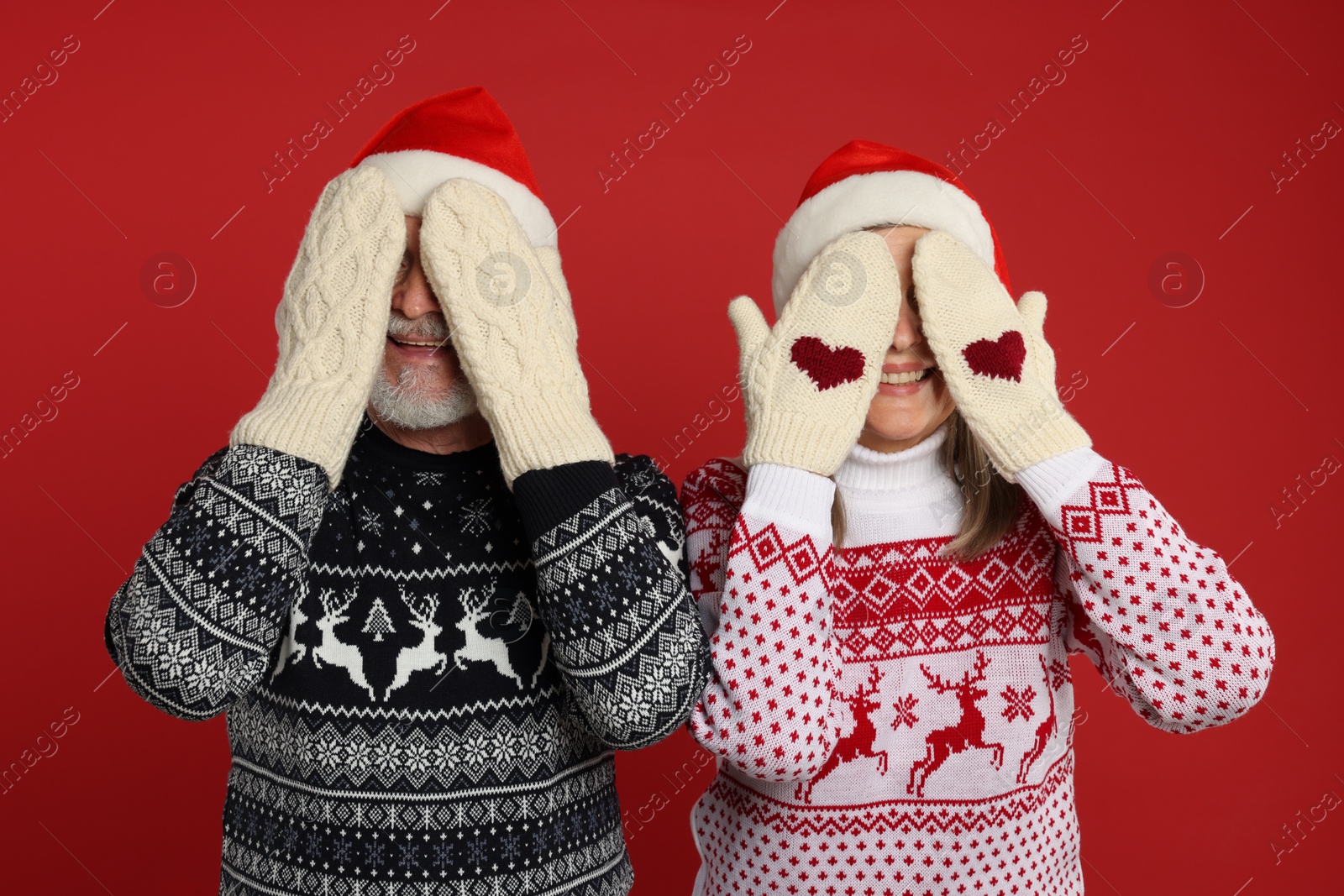 Photo of Senior couple in Christmas sweaters and Santa hats covering faces with hands in knitted mittens on red background