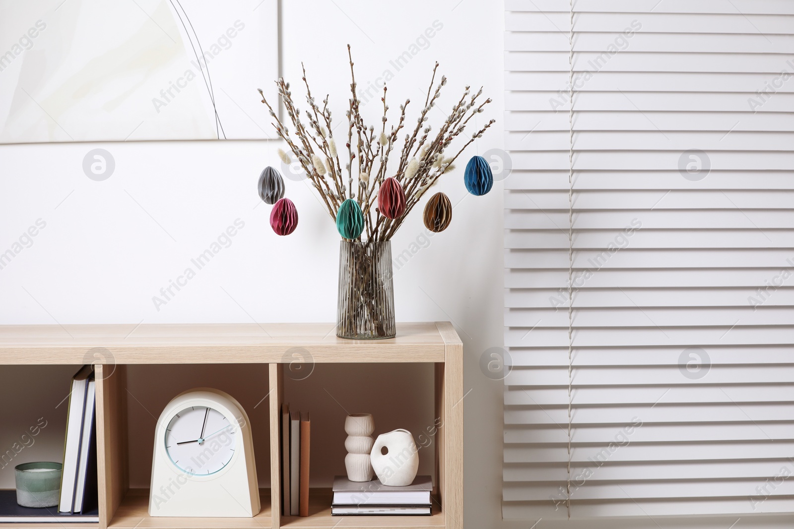 Photo of Beautiful pussy willow branches with paper eggs in vase on shelving unit at home. Easter decor