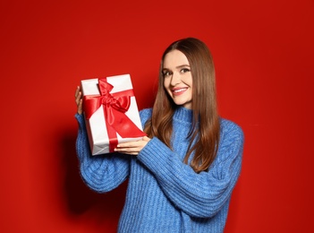 Young woman in Christmas sweater holding gift box on red background