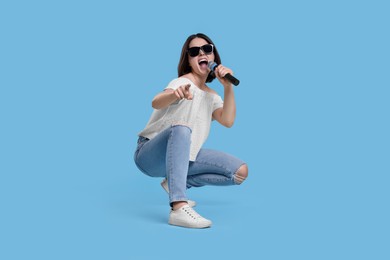 Beautiful young woman with sunglasses and microphone singing on light blue background