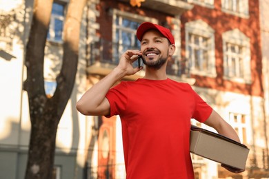 Photo of Courier with parcel talking on smartphone outdoors. Order delivery