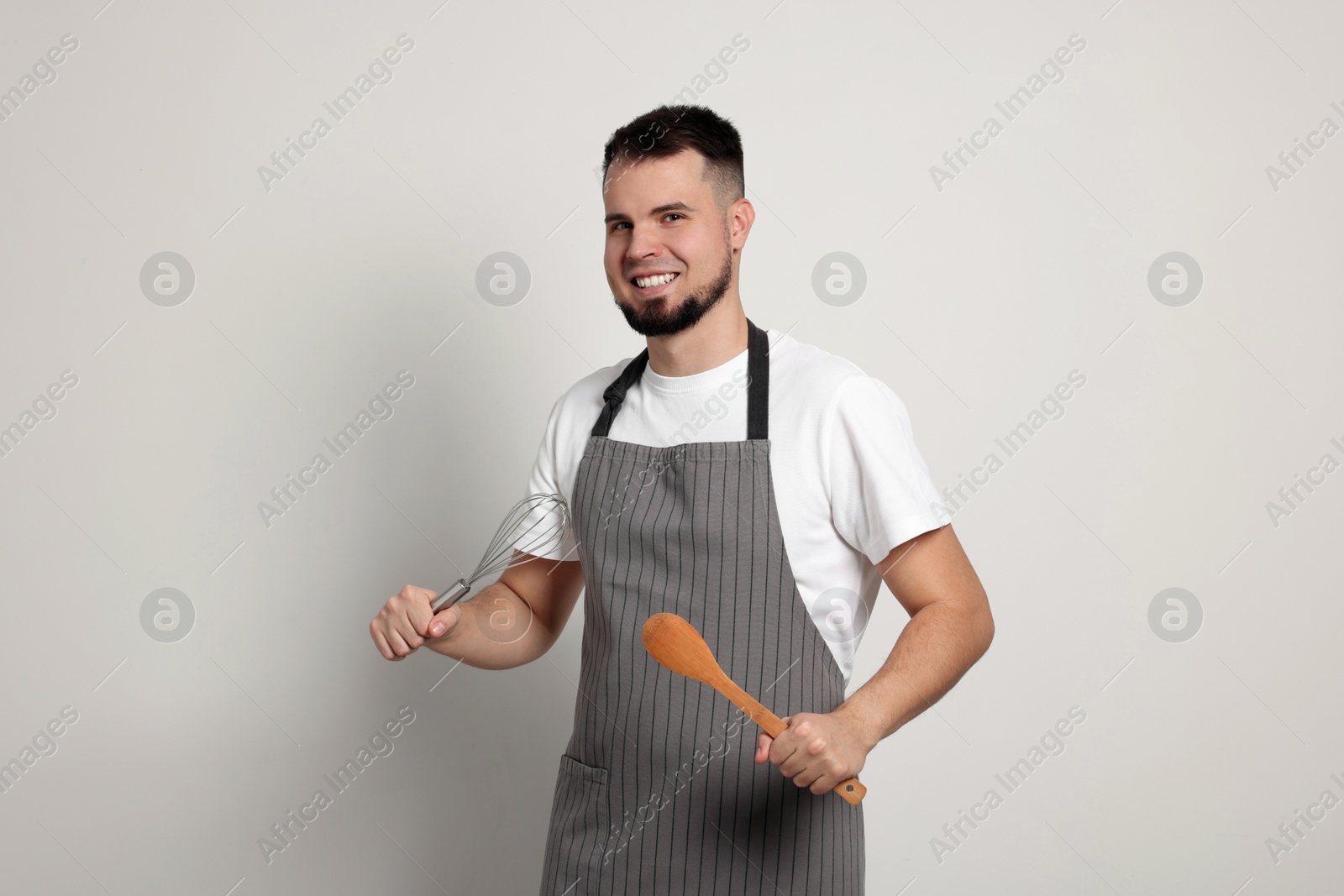 Photo of Happy professional confectioner in apron holding whisk and spoon on light grey background