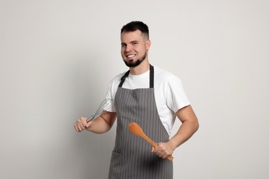 Happy professional confectioner in apron holding whisk and spoon on light grey background