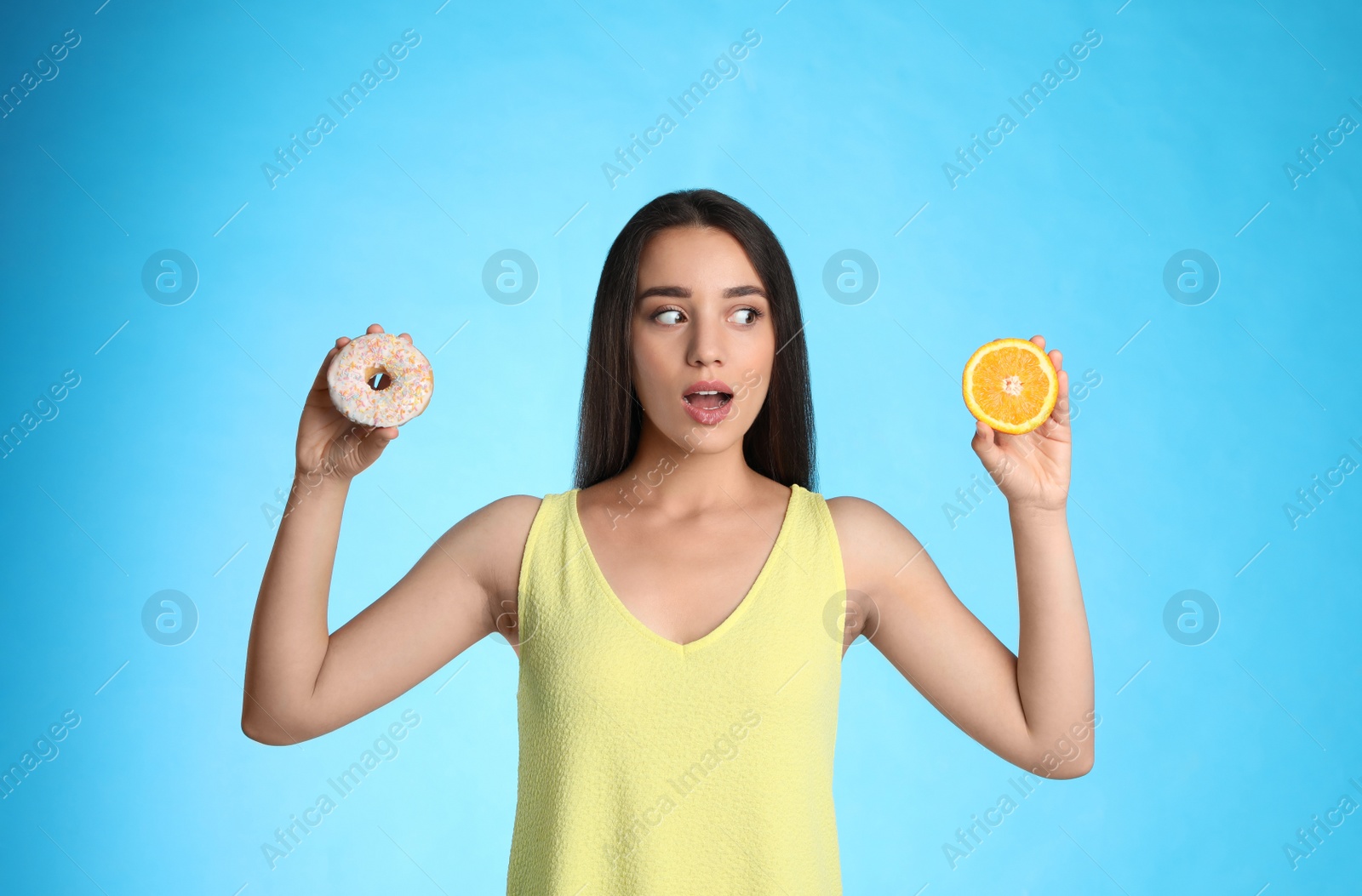 Photo of Woman choosing between orange and doughnut on light blue background