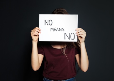 Photo of Young woman holding card with words NO MEANS NO against dark background