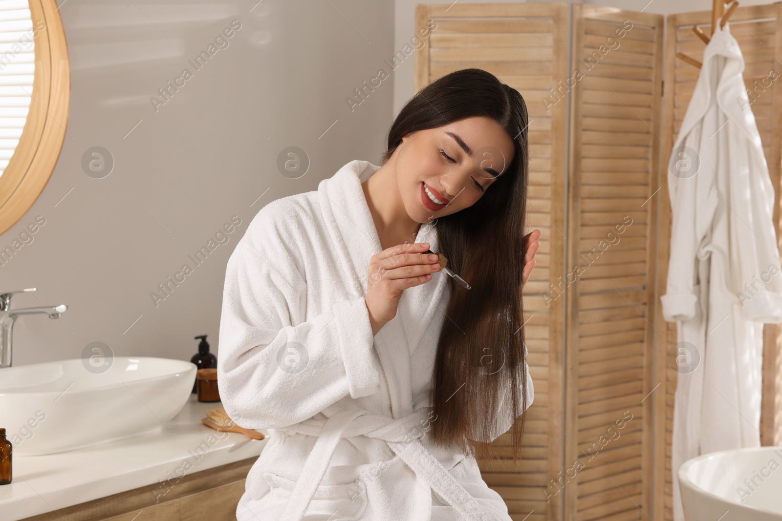 Photo of Happy young woman applying essential oil onto hair in bathroom