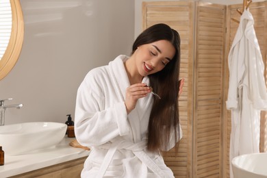 Happy young woman applying essential oil onto hair in bathroom